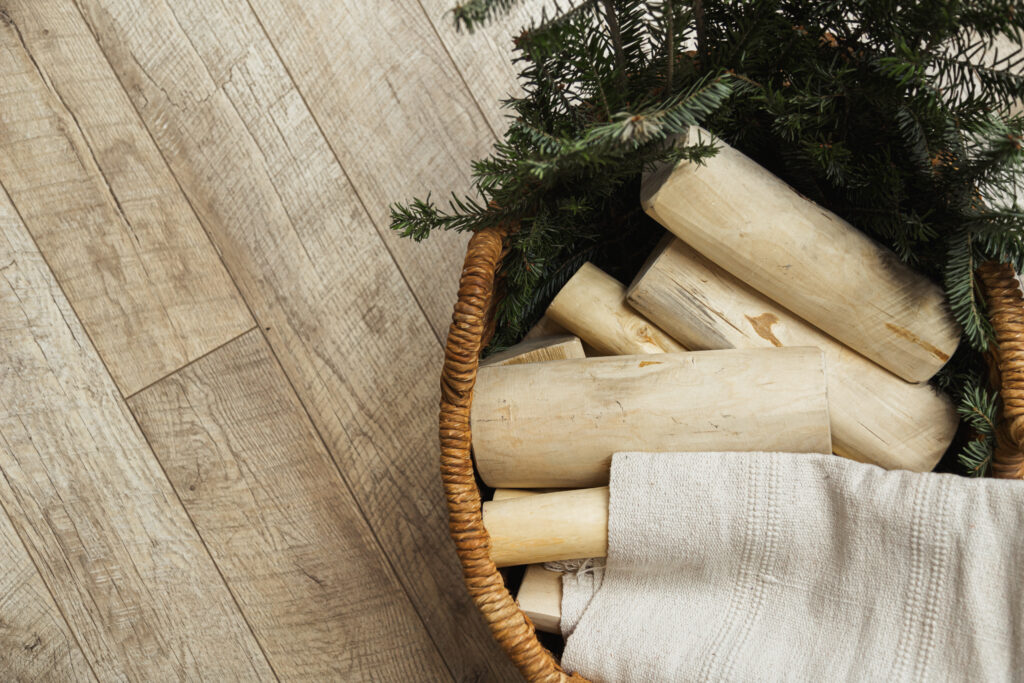 Basket with light birch logs and green pine branches, resting on a weathered wooden floor; a beige towel peeks from the basket edges.